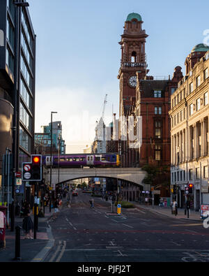Manchester, Inglaterra, Reino Unido - 30 de junio de 2018: una clase de ferrocarril norte 323 cruces de tren eléctrico de Manchester, Oxford Road, con la torre del reloj de t