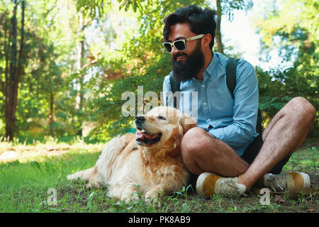 El hombre con la barba y su pequeño perro amarillo jugando y disfrutando del sol, la naturaleza y la amistad. Hombre Barbado en un cielo azul, camiseta y gafas de sol taping Foto de stock