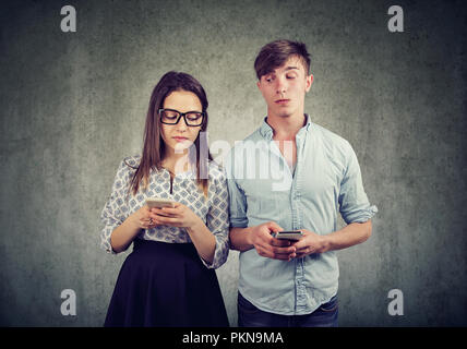 Joven Hombre y mujer mirando y peeking en smartphones de uno al otro con incredulidad