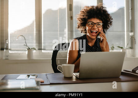 Retrato de la risa joven sentada en su escritorio en la oficina. La empresaria africanos hablando por teléfono móvil y riendo.