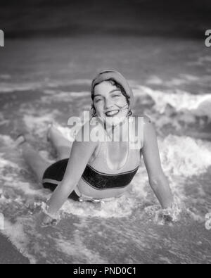 1920 MUJER MORENA sonriente tumbado en la playa, surfear olas llevar gorro de baño bañador mirando a la Cámara - b5831 HAR001 HARS SEÑORAS PERSONAS SURF B&W CONTACTO OCULAR MORENA SHORE felicidad ángulo alto alegre entusiasmo estilos de playas de recreo sonrisas alegres elegante traje de baño MODAS SEASHORE adulto joven mujer EN BLANCO Y NEGRO la etnia CAUCÁSICA HAR costera001 ANTICUADO