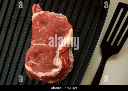 Un pedazo de carne cruda (ripe steak) sobre una parrilla eléctrica negra en un cuadro gris. Foto de stock
