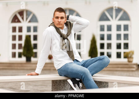 Elegante joven apuesto hombre de éxito en una camisa blanca con pantalones  de mezclilla sentado en un banco en el exterior del hotel Fotografía de  stock - Alamy