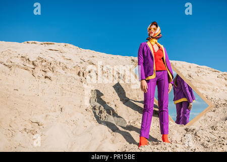 Hermoso modelo con ropas de colores posando cerca de espejo en una duna de arena con el cielo azul Foto de stock