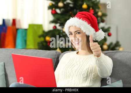 Mujer con un portátil y pulgares para arriba en navidad sentados en un sofá en el salón en casa Foto de stock
