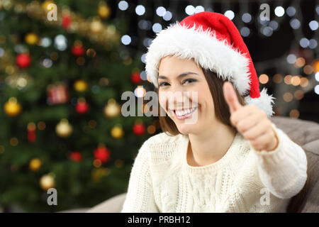 Feliz mujer con pulgares hacia arriba mirando en la navidad sentados en un sofá en el salón en casa Foto de stock
