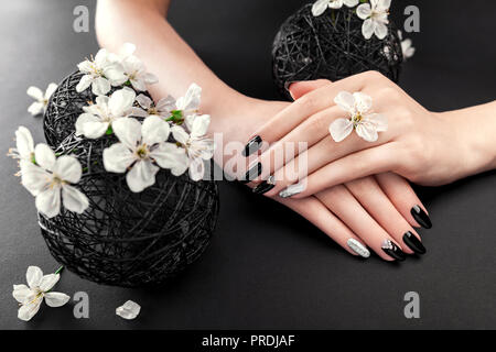 Negro y plata de manicura con los cerezos en flor sobre fondo negro. Mujer con uñas negras rodeadas con flores blancas. Cuidado del cuerpo