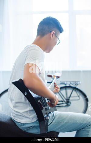Vista lateral del hombre asiático tocando la guitarra eléctrica desenchufada en casa