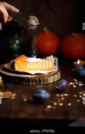 Lado femenino espolvorea azúcar en polvo sobre el cheesecake de calabaza. Calabazas, lámpara de mesa, follaje, madera de vainilla sobre un fondo oscuro. Foto de stock