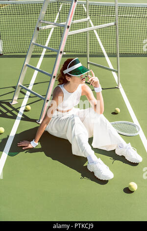 Moda joven en ropa blanca y tapa sentado en la pista de tenis con raquetas y pelotas. Foto de stock