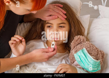 Una niña con su osito de peluche favorito a quien llevaba un vendaje de gasa está enfermo en la cama y su madre mide la temperatura con una elec Foto de stock