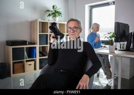 Hombre senior con cámara sentado en la oficina con su colega trabajando detrás de él Foto de stock