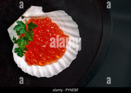 Rojo Salmón caviar en concha de vieira sobre una placa redonda de madera oscura con hierbas frescas, vista superior Foto de stock