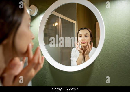 Mujer en albornoz haciendo un masaje facial mirando en el espejo redondo en el baño