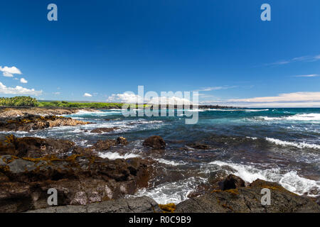 Vista al mar en la playa de arena negra en la isla más grande de Hawai. Océano Pacífico azul con olas, áspera roca volcánica y vegetación marrón en la orilla.
