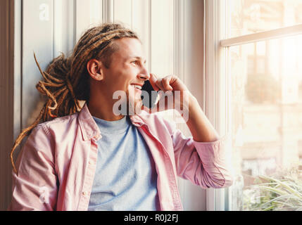Hipster atractivo hombre barbado con temores usando audífonos sentado cerca de la ventana y hablando sobre el smartphone con su mejor amigo Foto de stock