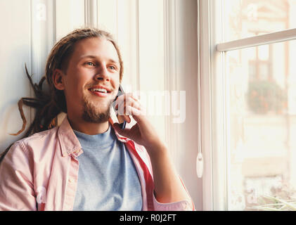Hipster atractivo hombre barbado con temores usando audífonos sentado cerca de la ventana y utilizando el smartphone Foto de stock