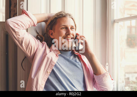 Hipster atractivo hombre barbado con temores usando audífonos sentado cerca de la ventana y la celebración de smartphone, él habla con su amigo Foto de stock