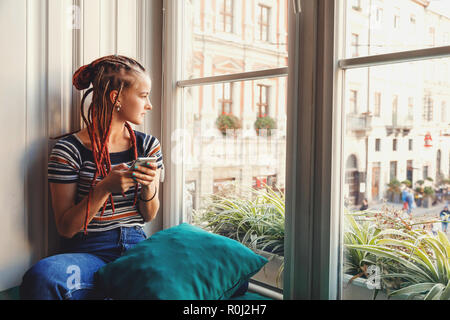 Hipster Estudiante lindo chica con temores mirando por la ventana y pensando en sus amigos mientras escribiendo el mensaje en el smartphone Foto de stock
