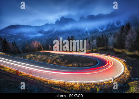 Los faros de un coche borrosa en la sinuosa carretera de montaña con nubes bajas por la noche en el otoño. Espectacular paisaje con camino asfaltado, estelas de luz, foggy f Foto de stock