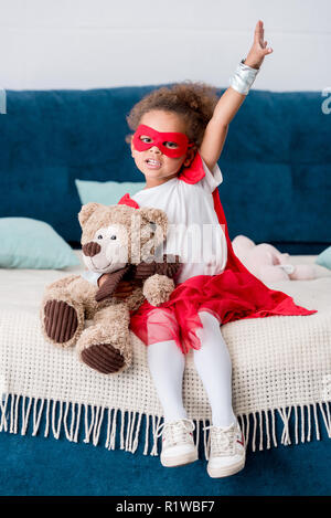 Adorable pequeño niño africano-americanos en traje de superhéroe sentada en la cama con oso de peluche