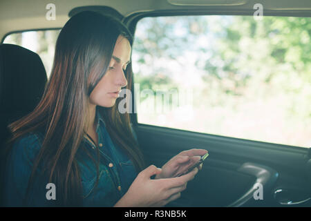 Hermosa mujer sonriente mientras está sentado en el asiento del acompañante en el coche. Chica está utilizando un smartphone
