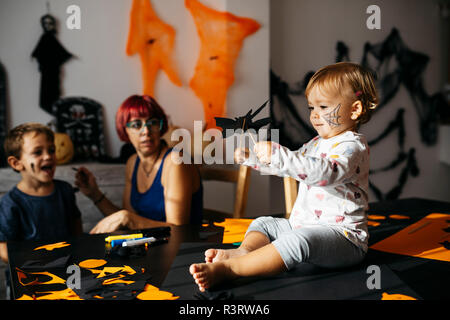 Bebé niña jugando con decoración de Halloween en casa mientras observando a su madre y hermano