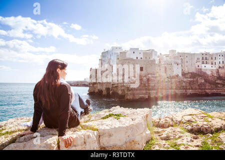 Italia, Apulia, Polognano a Mare, vista posterior de la mujer relajante en las rocas mirando el histórico casco antiguo