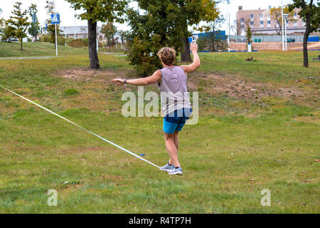 Un hombre joven en un parque público aprendiendo a caminar sobre la cuerda floja. Slacklining es una práctica de equilibrio que normalmente utiliza tensión cincha poliéster o nylon Foto de stock