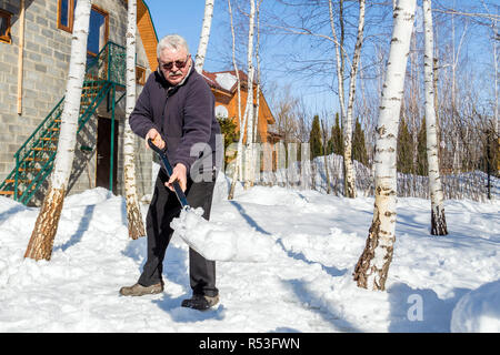Altos hombre arrojar nieve con pala de casa privada patio en invierno en día soleado. Anciano retirar nieve en el jardín tras fuertes snowfal Foto de stock