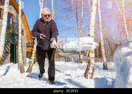 Altos hombre arrojar nieve con pala de casa privada patio en invierno en día soleado. Anciano retirar nieve en el jardín tras fuertes snowfal Foto de stock