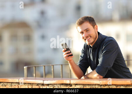 Feliz Hombre sujetando un teléfono inteligente mirando a la cámara en un balcón al atardecer