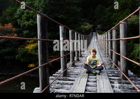 Un muchacho joven con camiseta amarilla sentado y meditando con los ojos cerrados en un puente colgante de madera, Wakayama, Japón