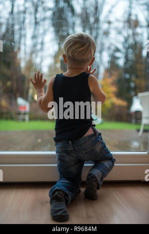 Niño chico de pie junto a una ventana, mirando hacia fuera para observar la naturaleza. Foto de stock
