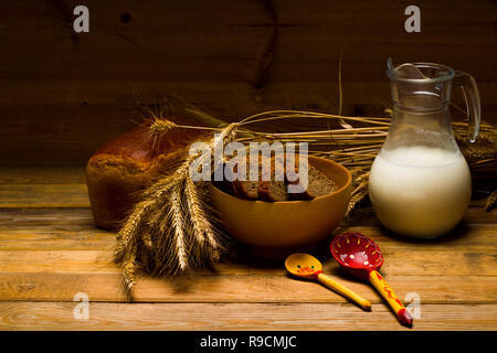 Bodegón con una jarra de cristal de leche, una jarra de cristal, el pan de centeno, mazorcas de maíz en el fondo de los tableros de madera Foto de stock