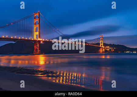 La vista nocturna del Puente Golden Gate se refleja en la superficie de agua borrosa de la bahía de San Francisco, fondo de cielo azul oscuro; California