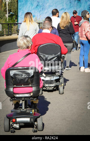 Vista trasera de un hombre y una mujer, ambos en silla de ruedas, scooters estilo unirse a las multitudes en la acera mientras turismo en los EE.UU. Foto de stock