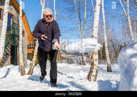 Altos hombre arrojar nieve con pala de casa privada patio en invierno en día soleado. Anciano retirar nieve en el jardín tras fuertes snowfal Foto de stock