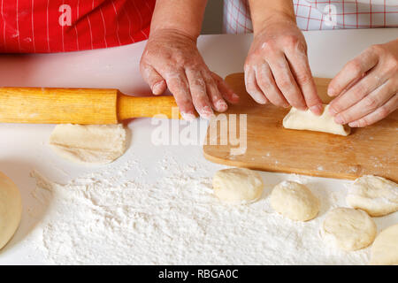 Close-up. La mujer de edad en el delantal de cocina rojo desenrolla la masa con un rodillo, la hija adulta esculpe tartas Foto de stock