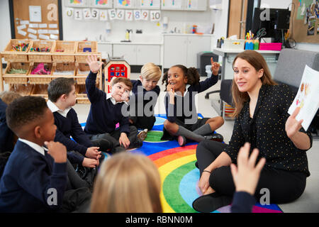 La escuela primaria los niños sentados en el suelo en un aula con su profesor, levantando las manos para contestar a una pregunta, el enfoque selectivo Foto de stock