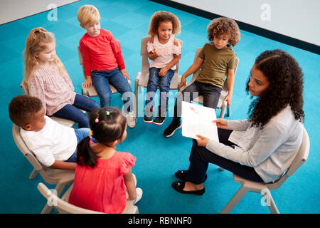 Profesor de la escuela femenina joven leyendo un libro a los niños de la escuela infantil, sentados en sillas en un círculo en el aula escuchando, niveles elevados de ver Foto de stock
