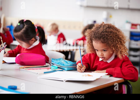 Colegiala vistiendo uniforme escolar sentado en una mesa en un aula de la escuela infantil de dibujo, cerrar Foto de stock