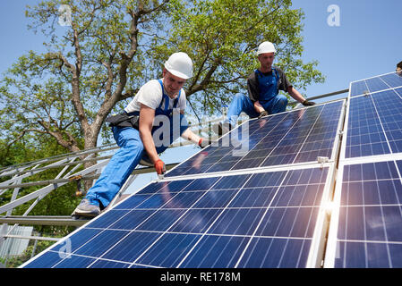 Dos jóvenes técnicos instalando pesados fotovoltaicos solar panel sobre plataforma de acero alto en árbol verde de fondo. Panel solar exterior Voltaicas sistema instalación, trabajo peligroso concepto. Foto de stock
