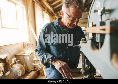 Senior masculino carpintero trabajando en su taller. Carpenter cortando madera en la máquina en el taller de carpintería. Foto de stock