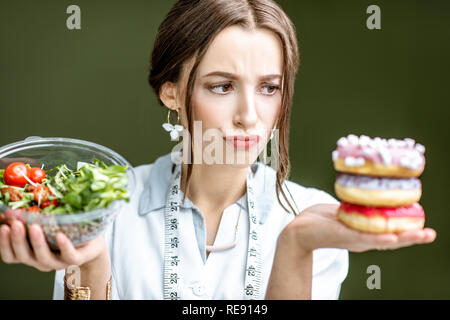 Mujer joven nutricionista busca en las donas con tristes emociones elegir entre ensalada y postre insalubres en el fondo verde