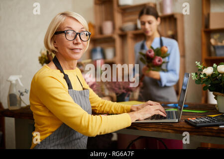 Vista lateral retrato de mujer madura dueño de la pequeña empresa con ordenador portátil y mirando a la cámara en la floristería, espacio de copia