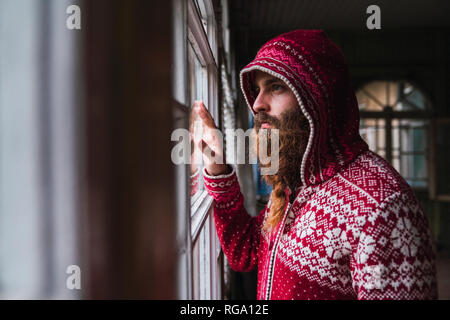 Retrato del hombre pensativo con barba vistiendo chaqueta con capucha mirando afuera de la ventana