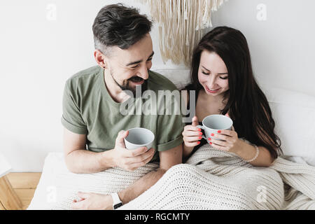 Feliz pareja sentada en la cama, beber café, divertirse