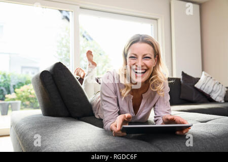 Retrato de mujer de risa tumbado en sofá en casa utilizando tablet Foto de stock