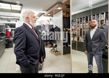 Los ancianos, hombre barbado con cabello gris tratando el clásico traje con la corbata y la camisa en la boutique. Hombre mirando en el espejo y ver la reflexión de los jóvenes a la moda y hombre encantador. Caro prenda de ropa de hombre.